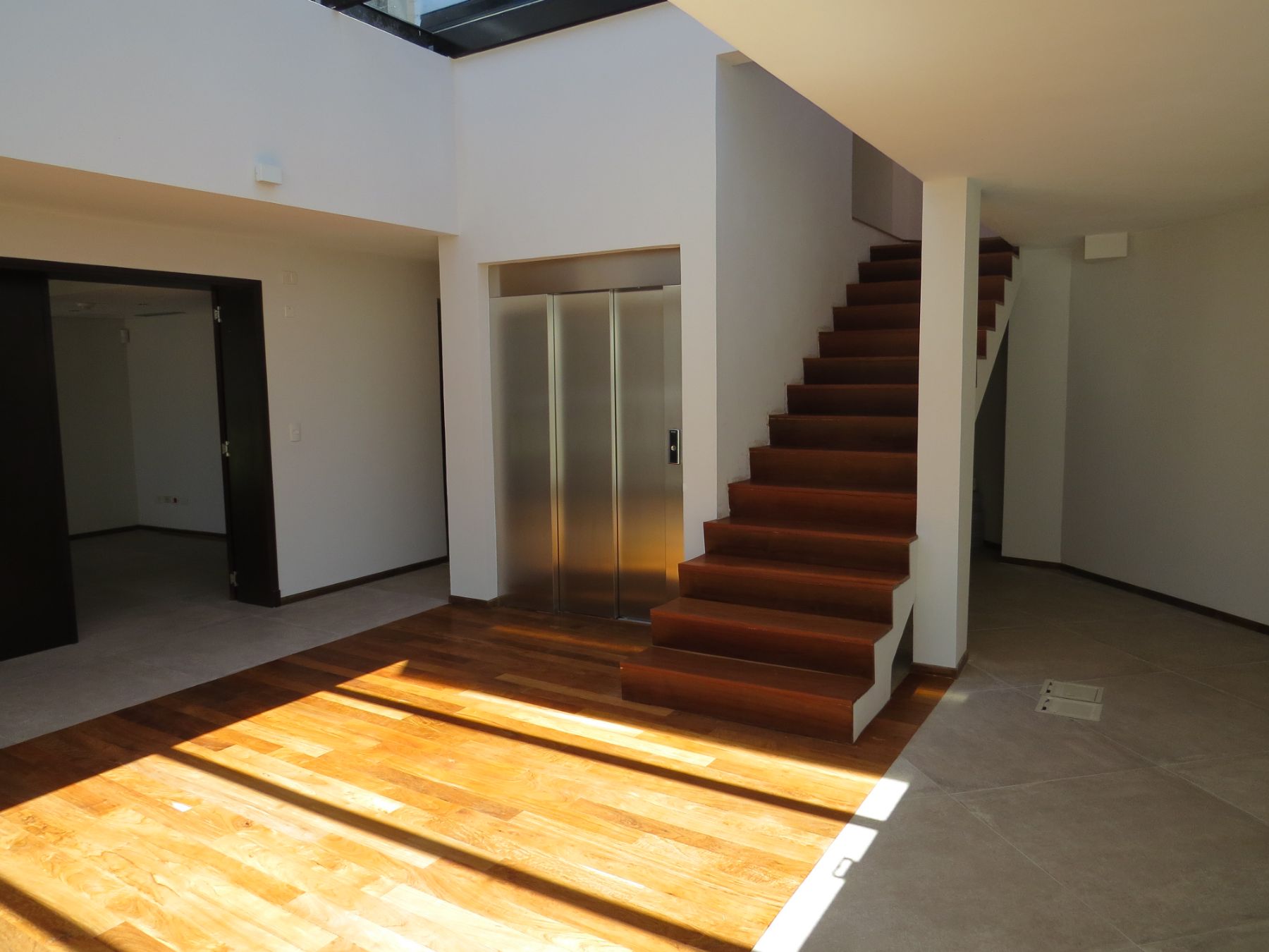 En la foto se aprecia el recibidor de la clínica de punta del este. El mismo posee un piso de madera en el centro y porcelanato hacia los costados. La madera da a su vez paso a la escalera de madera y el ascensor del edificio, el cual es un ascensor de acero inoxidable.